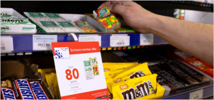 Person grabbing multi-colored Tic Tacs from shelf with banner displaying Exxon Mobil Rewards+ gas rewards points offer of 80 points